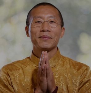 Master Mingtong Gu, wearing a golden embroidered traditional Chinese garment, is pictured with his hands gently pressed together in a gesture of gratitude or prayer. He has a warm, serene smile, and his expression radiates calmness and compassion. The softly blurred background suggests a natural, light-filled setting.