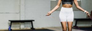 A cropped image of a fit woman in motion, jumping rope in a minimalistic indoor gym setting. She wears a black sports top and white high-waisted shorts, with her arms extended as she swings the jump rope. The background is softly blurred, focusing attention on her form and movement, capturing the energy and intensity of the workout.
