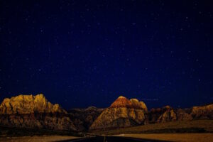 A breathtaking view of rugged desert mountains illuminated under a vast, starry night sky. The road in the foreground leads into the serene landscape, inviting a sense of wonder, solitude, and connection with the universe.