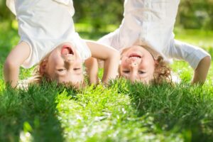 wo cheerful children playing outdoors, both upside down with their heads on the grass and legs in the air. They are laughing and enjoying the sunny day, surrounded by vibrant green grass and a bright natural setting.