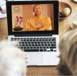 A laptop screen displaying a Zoom session with Master Mingtong Gu wearing a golden traditional outfit, offering a warm, heartfelt gesture with his hand on his chest.