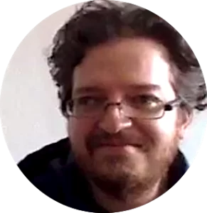 Portrait of a man with curly hair and glasses, smiling softly, set against a neutral indoor background, displayed within a circular frame.