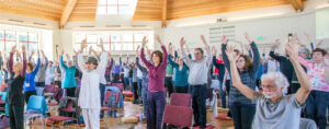 Group of individuals participating in a guided Qigong practice session indoors, raising their arms in unison under natural light streaming through large windows.