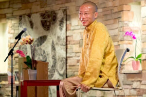 Master Mingtong Gu smiling warmly while teaching a session on Wisdom Healing Qigong, seated in a serene setting with vibrant orchids and a stone wall backdrop.
