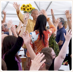 Master Mingtong Gu leading a vibrant Qigong session in a dome setting, surrounded by participants raising their hands, with bright sunflowers in the background adding a touch of nature and warmth.