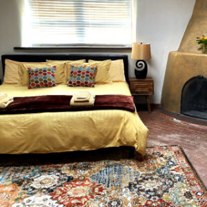 Cozy and inviting bedroom with a warm yellow bedspread, colorful accent pillows, a patterned area rug, and a traditional kiva fireplace, creating a blend of comfort and Southwestern charm.