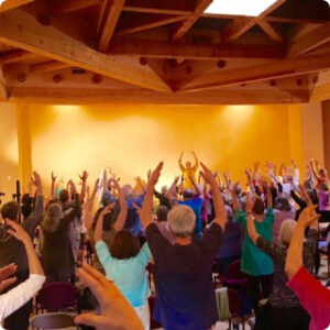 Group of individuals practicing Qigong in a spacious room with a warm, golden ambiance, guided by an instructor under a wooden beam ceiling.