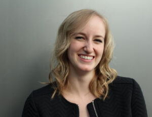 Portrait of a smiling woman with blonde wavy hair, wearing a black textured jacket against a neutral gray background, conveying professionalism and approachability.