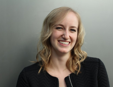 Portrait of a smiling woman with blonde wavy hair, wearing a black textured jacket against a neutral gray background, conveying professionalism and approachability.
