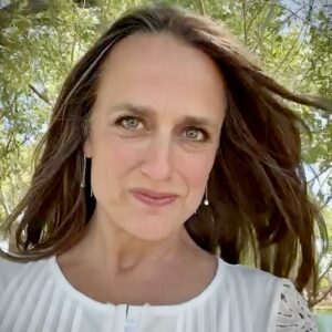 Portrait of Sarah, smiling outdoors with flowing hair and a natural background of greenery and sunlight, wearing a white blouse and drop earrings.