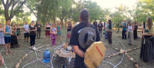 A sacred outdoor ceremony with a diverse group of people gathered in a circle around a central altar, featuring stones and symbolic items, set in a lush, tree-filled environment.