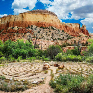 A vibrant labyrinth in the foreground set against the stunning red rock cliffs and greenery of Ghost Ranch in New Mexico under a partly cloudy sky.