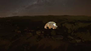 A nighttime aerial view of the Chi Center Dome glowing beautifully under a star-filled sky, surrounded by a tranquil landscape of trees and adobe-style buildings