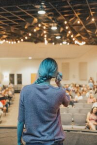 A person with blue hair, seen from behind, is standing on a stage in a softly lit room with string lights hanging from the ceiling.