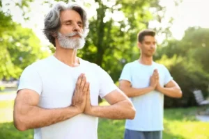Two men practicing mindfulness and meditation outdoors, standing in a serene park with hands in prayer position, surrounded by lush greenery and soft sunlight.