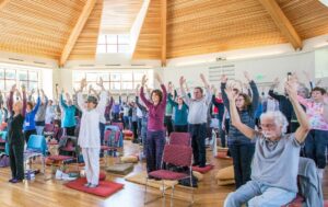 Large group of people practicing Qigong indoors in a bright, spacious room with wooden ceilings and natural light, raising their arms in unison for a guided energy movement session.