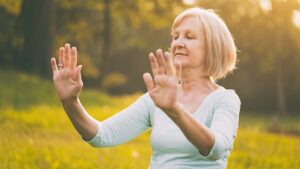 Senior woman practicing Qigong in a serene outdoor setting, with closed eyes and graceful hand movements, surrounded by warm sunlight and lush greenery.