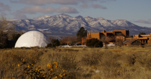 Stunning view of The Chi Center Dome and adobe-style buildings set against the backdrop of snow-capped mountains, surrounded by natural desert landscape and clear skies.