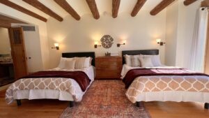 Cozy and elegantly designed bedroom at The Pueblo House, featuring two twin beds with plush bedding, a rustic wooden dresser, warm lighting, and exposed wooden beams for a serene and inviting retreat experience.