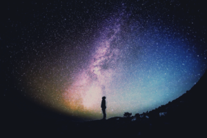 Silhouette of a person standing on a hill, gazing at a breathtaking view of the Milky Way galaxy, with vibrant hues of purple, blue, and gold illuminating the night sky.