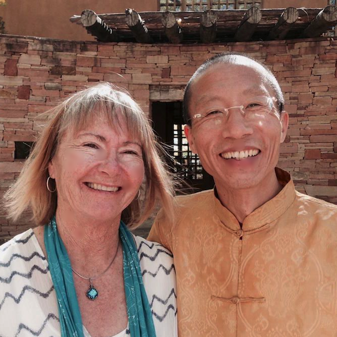 Master Mingtong Gu smiling alongside a woman in front of a rustic stone structure, symbolizing connection, joy, and healing energy.