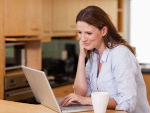 Smiling woman working on a laptop in a modern kitchen setting with a coffee mug on the counter, symbolizing a comfortable and productive home workspace.
