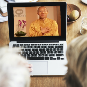 Master Mingtong Gu appearing on a laptop screen during a Zoom session, wearing a golden traditional outfit and holding his hand to his heart, symbolizing connection and compassion during an online Wisdom Healing Qigong event.