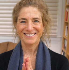 A smiling woman with hands in a prayer position, exuding warmth and compassion, standing in a cozy room with bookshelves in the background, symbolizing mindfulness and gratitude.
