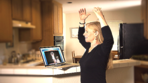 A woman practicing Qigong at home while following an online session on her laptop, raising her arms in a peaceful kitchen environment, illustrating accessible virtual wellness practices.
