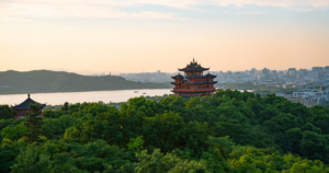 Traditional Chinese pagoda surrounded by lush greenery overlooking a serene river and a modern cityscape under a pastel-colored sunset sky.