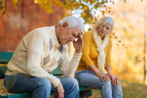Senior man sitting on a bench in distress, holding his head, while an empathetic woman looks on in concern, surrounded by autumn foliage.