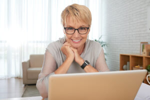Smiling senior woman with short blonde hair wearing glasses, seated in a bright living room, looking at a laptop, symbolizing engagement, learning, and modern connectivity.