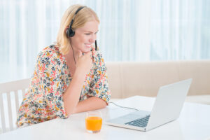 Young woman with blonde hair wearing a floral blouse and headphones, smiling while engaging in an online meeting on her laptop, with a glass of orange juice on the table, symbolizing focus, learning, and virtual connectivity.