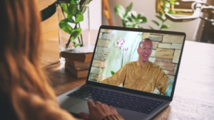 Person watching a video of Qigong Master Mingtong Gu in a golden robe, smiling against a stone wall backdrop, with plants and books on a wooden desk, representing online wellness learning.