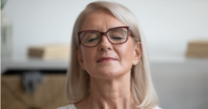 A serene older woman with glasses and silver hair, sitting with her eyes closed in a calm indoor setting, symbolizing mindfulness, relaxation, and inner peace.