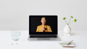 A laptop displaying Qigong Master Mingtong Gu in a golden robe on a minimalist desk with a vase, notebook, and glass of water, symbolizing mindfulness and online learning.