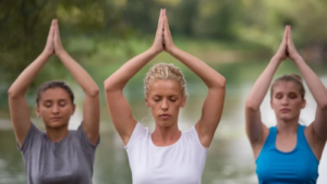 Three women practicing mindfulness and meditation outdoors near a serene lake, focusing on inner peace and relaxation.