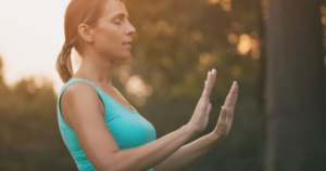 A woman practicing Qigong in an outdoor setting during sunset, wearing a turquoise tank top, with a peaceful expression and her hands extended in a meditative posture, symbolizing mindfulness and connection to nature.
