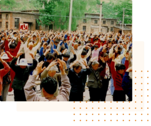 Large group of people practicing Qigong outdoors in a community setting, performing synchronized movements with raised arms. The gathering takes place in a rural village surrounded by traditional buildings and lush greenery.