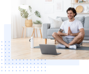 Young man practicing mindfulness at home, sitting cross-legged on a yoga mat with hands in a prayer position, smiling at his laptop during an online meditation session. The cozy living room features bright natural light, a modern gray sofa, and greenery.