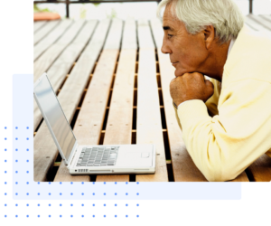 An older man with white hair intently watching his laptop screen while sitting outdoors on a wooden deck, representing learning, connection, and engagement in a serene environment.