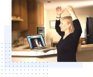 A woman practicing Qigong at home while following an online session on her laptop, raising her arms in a peaceful kitchen environment, illustrating accessible virtual wellness practices.