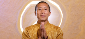 Master Mingtong Gu wearing a golden traditional outfit with hands in a prayer gesture, smiling warmly against a glowing circular sand-inspired background.
