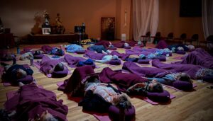 Participants lying on mats and cushions during a Kiva healing session, covered with blankets in a serene, softly lit room with Master Mingtong Gu guiding the session from the stage.