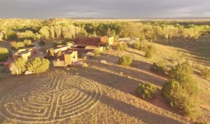 Aerial view of The Chi Center in Galisteo, New Mexico, featuring traditional adobe architecture surrounded by natural landscapes, trees, and a labyrinth design in the foreground under a golden sunset.