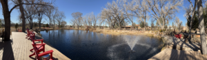 A panoramic view of The Chi Center's tranquil koi pond surrounded by trees and red Adirondack chairs, with a fountain creating ripples under the bright blue sky in Galisteo, New Mexico.