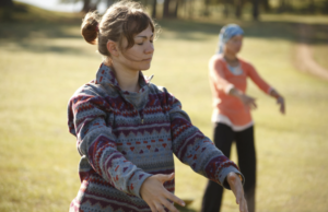 Two women practicing Qigong outdoors in a sunlit grassy field, focusing on gentle movements and mindfulness, with a serene and peaceful atmosphere.