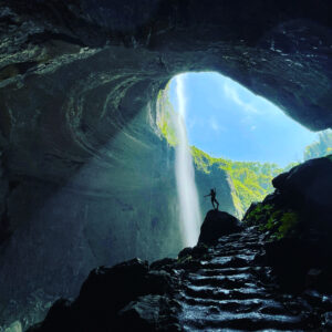 Silhouette of a person standing triumphantly in a stunning cave with sunlight streaming through the entrance and a waterfall in the background, symbolizing adventure and connection with nature.