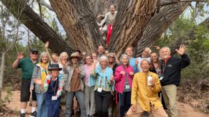 Master Mingtong Gu with a group of joyful participants gathered around the grandmother tree, sharing smiles and energy in a serene natural setting.