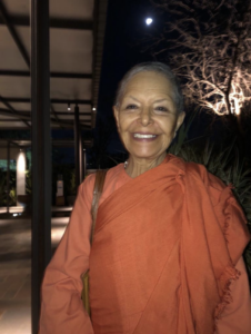 Smiling woman in an orange traditional robe, standing outdoors under the moonlight, with softly illuminated trees and modern architectural elements in the background.